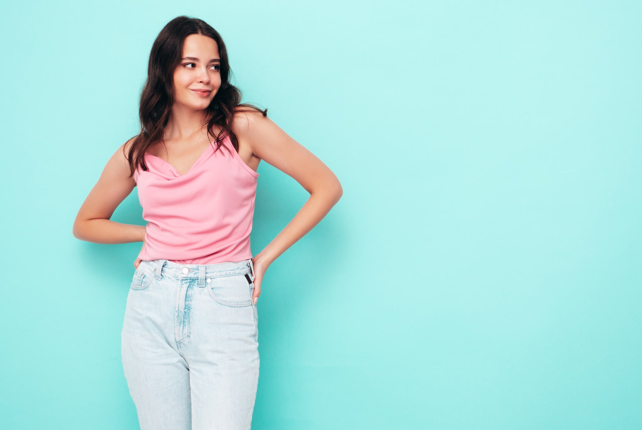 A woman with a Rectangle Body Shape in a pink sleeveless top and light blue jeans stands against a turquoise background, looking to the side with a slight smile.